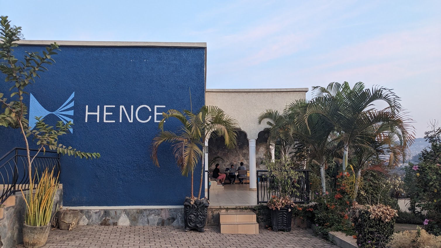 The Hence office in Kigali, Rwanda — a navy building with the Hence mural, surrounded by palm trees, with team members working under an open arch.